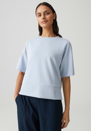 Frau trägt ein hellblaues kurzärmeliges Oberteil und marineblaue Hose, steht vor einem schlichten hellgrauen Hintergrund.