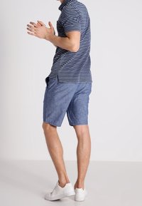 Homme portant un polo rayé bleu, un short bleu et des baskets blanches, se tenant les mains jointes devant un fond uni.