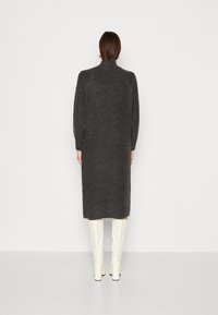 Donna in piedi con le spalle rivolte verso l'osservatore, che indossa un lungo abito di maglia grigio scuro con maniche lunghe e stivali bianchi sopra il ginocchio, su uno sfondo bianco semplice.