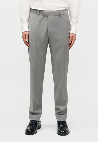 Άνδρας που φοράει ανοιχτό γκρι παντελόνι φόρμας, μαύρα δερμάτινα παπούτσια και λευκό πουκάμισο με μακριά μανίκια, στέκεται μπροστά σε απλό φόντο.