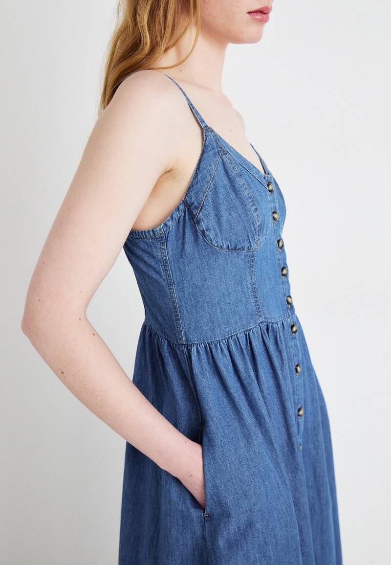 Robe en denim avec un corsage ajusté, des bretelles spaghetti, un devant à boutons et une jupe fluide. Elle comprend des poches latérales et est de couleur bleu clair.