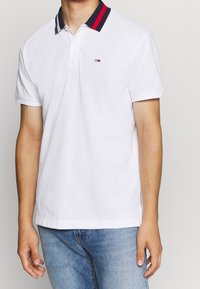 Bílá polo košile s dvoubarevným límcem v červené a navy pruhované kombinaci. Na levé hrudi je malé značkové logo. Materiál: bavlna.