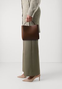 Brun strukturerad handväska med ett handtag, bärs tillsammans med en ljusgrå stickad cardigan och olivgröna vida byxor, i sällskap av beige klackar.