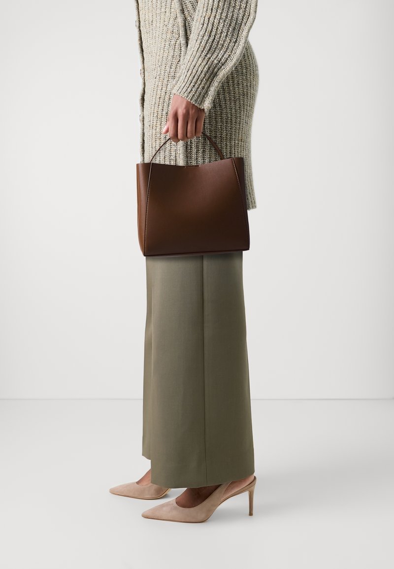 Brun strukturerad handväska med ett handtag, bärs tillsammans med en ljusgrå stickad cardigan och olivgröna vida byxor, i sällskap av beige klackar.