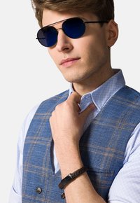 Okrągłe okulary przeciwsłoneczne z ciemnoniebieskimi soczewkami, niebiesko-brązowa kamizelka w kratę oraz jasnoniebieska koszula w wzory. Na nadgarstku wpleciona brązowa bransoletka.