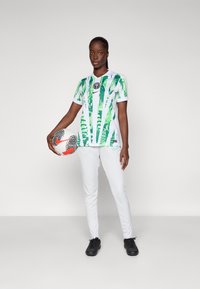 Žalias ir baltas futbolo marškinėliai su lapų raštu, juoda emblema, balti kelnių, ir juodi batai. Žaidėjas laiko raštuotą futbolo kamuolį.