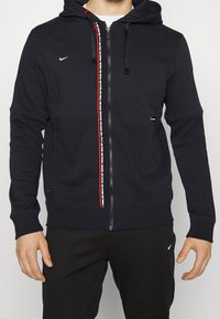 Sweat à capuche noir avec une fermeture éclair à l'avant, des accents rouges, un logo blanc et une bande de marque sur la fermeture éclair. Comprend une capuche avec cordon de serrage et des poignets côtelés.