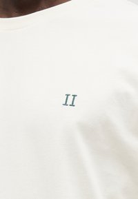 Μπεζ βαμβακερό t-shirt με στρογγυλή λαιμόκοψη, που διαθέτει μικρό πράσινο κεντημένο «II» στην αριστερή πλευρά του στήθους. Μαλακή υφή, casual σχεδιασμός.