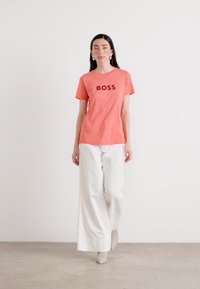 Modelis pārliecinoši iet, ģērbies korallkrāsas T-kreklā ar uzrakstu "BOSS" melnā krāsā, kas kombinēts ar baltām platiem biksēm un beige krāsas potītes zābakiem.
