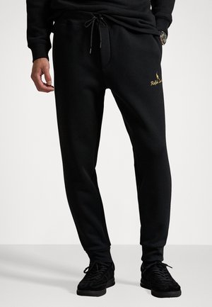 Mann trägt schwarze Jogginghose mit Kordelzug und goldenem Ralph-Lauren-Logo, kombiniert mit schwarzen Sneakern, steht vor weißem Hintergrund.