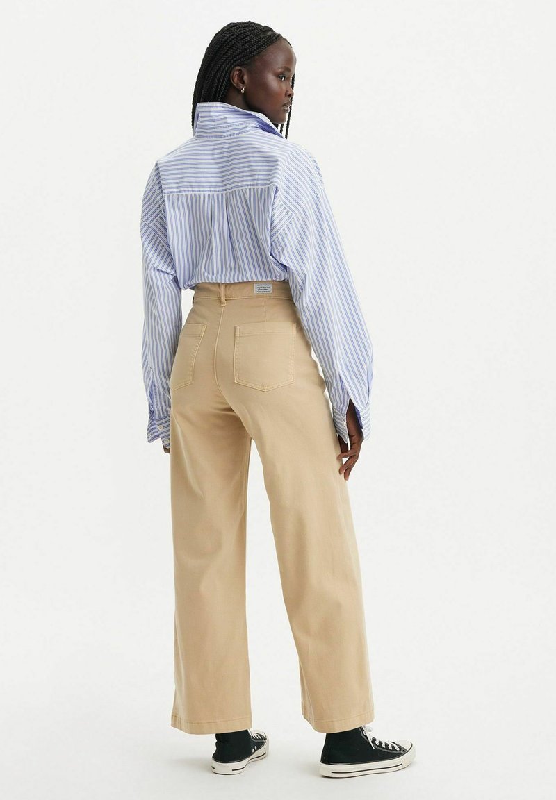 Camisa de manga larga a rayas azul y blanca, pantalones beige de pierna ancha y zapatillas negras de caña alta. Bolsillos traseros visibles en los pantalones.