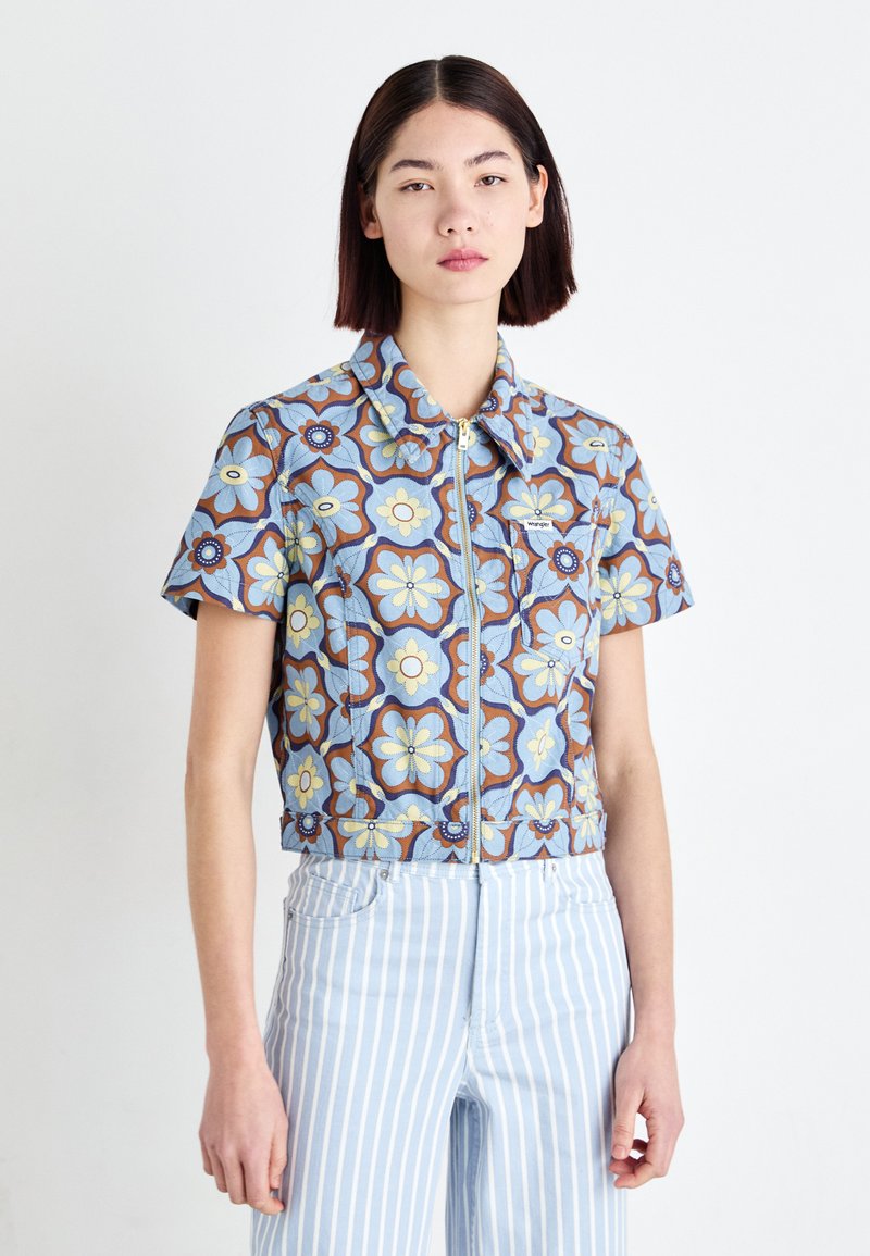 Kortermet crop-skjorte med et blått blomsterdesign og gule og brune detaljer. Den har en glidelås foran og en spiss krage.