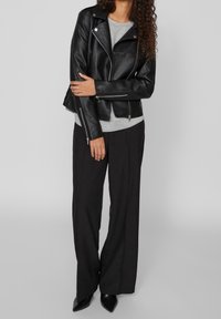 Veste de moto en cuir noir avec des garnitures en métal argenté, fermeture éclair à l'avant et coupe ajustée, portée sur une chemise grise et un pantalon large noir.