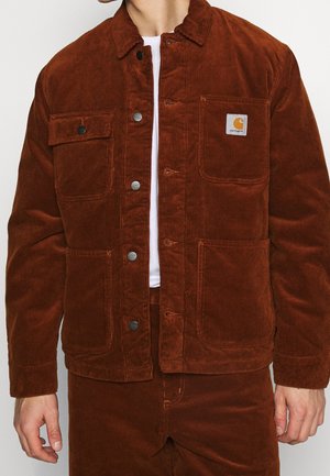 Mężczyzna w rdzawej kurtce sztruksowej z zapięciem na guziki, kieszeniami na piersi i naszywką z logo Carhartt na białej koszuli.