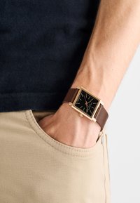 Négyzet alakú óra aranyszínű tokkal, fekete számlappal és barna bőr szíjjal. Vékony órajelzőkkel és piros másodpercmutatóval rendelkezik.