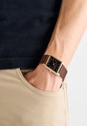 Orologio quadrato con cassa dorata, quadrante nero e cinturino in pelle marrone. Mostra sottili indici delle ore e una lancetta dei secondi rossa.