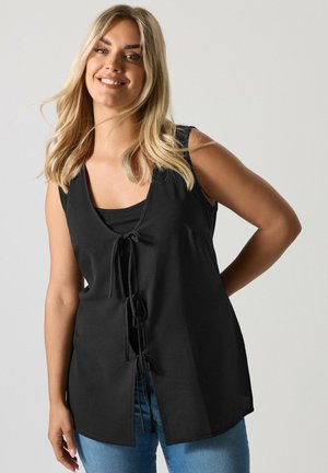 Donna bionda sorridente, indossa una canotta nera senza maniche con fiocchi frontali sopra una canottiera nera, abbinata a jeans blu, in piedi davanti a uno sfondo chiaro.