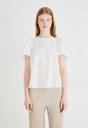 Hvit bluse med puffermer, rund halslinning og scallop-kant. Laget av lett materiale, brukt med strukturerte beige bukser.