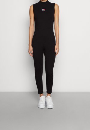 Person iført en sort ærmeløs tætsiddende jumpsuit med Tommy Jeans-logo og hvide plateau sneakers, stående mod en ensfarvet baggrund.