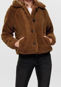Brun, fluffig fleecjacka med krage, fyra stora svarta knappar och två sidofickor, tillsammans med mörka jeans.