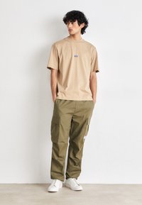 Beige t-shirt med et lille blåt logo, parret med olivengrønne cargo-bukser med flere lommer. Modellen bærer hvide sneakers.