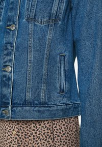 Denim jacket in medium blue, featuring silver buttons, two chest pockets, and a slightly cropped fit. The texture is rugged with visible stitching.