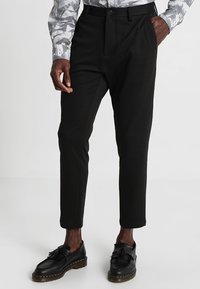 Pantalon noir sur mesure à jambe droite, avec un devant plat et sans poches apparentes, associé à des chaussures noires en cuir à pampilles.
