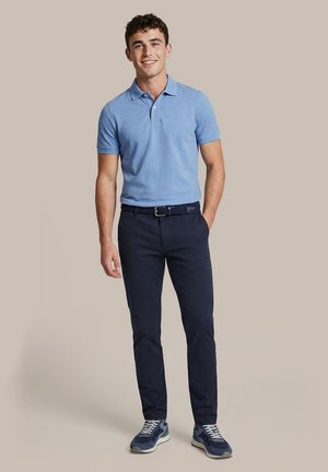 Jonge man staat, draagt een lichtblauw poloshirt, donkerblauwe broek met riem en donkerblauwe sneakers op een effen achtergrond.