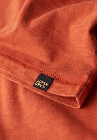 Roestkleurige stof met een gladde textuur, voorzien van een zwart "SUPER DRY" label met gouden letters op een gevouwen rand.