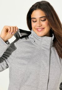 Chaqueta gris con cremallera y cuello alto, fabricada en un tejido texturizado, con cierres a presión y bolsillos ocultos. El interior presenta un material negro contrastante.