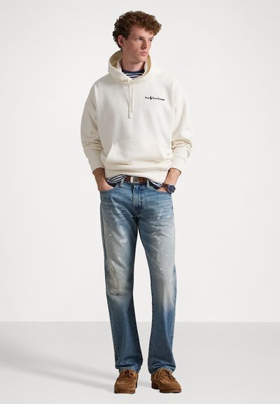 Κρέμ φούτερ με μπροστινή τσέπη και λογότυπο, συνδυασμένο με ανοιχτό μπλε τζιν με πιτσιλιές χρώματος και καφέ υπόδηση. Ορατές διακριτικές ρίγες.