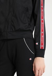 Svart idrottsjacka med dragkedja, framfickor och röd sidostripa med logotyp. Kombinerad med svarta byxor och vita kantpipningar.