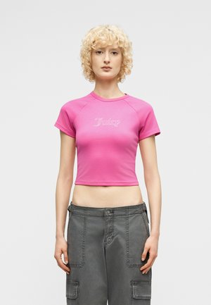 Ξανθός/ιά που φοράει ροζ κοντό T-shirt με το κείμενο «Juicy» και φαρδιά γκρι παντελόνια cargo, στέκεται μπροστά σε ένα απλό φόντο.