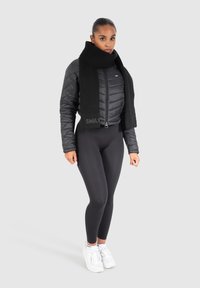 Black puffer jacket with quilted design, featuring a high collar, paired with a knit black scarf. Model wears black leggings and white sneakers.