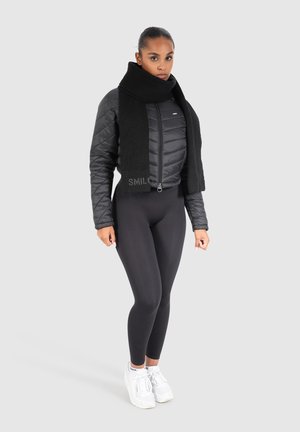 Sort pufferjakke med quiltet design, med høj krave, parret med et strikket sort tørklæde. Modellen har sorte leggings og hvide sneakers på.