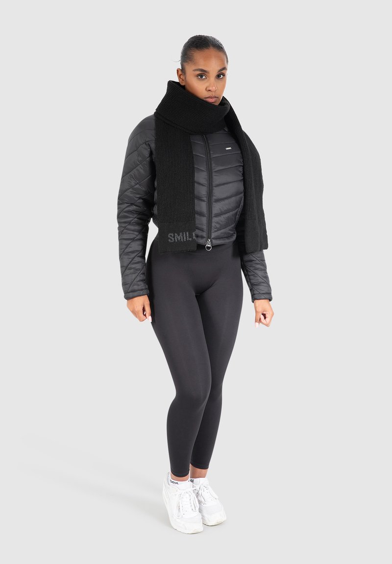 Black puffer jacket with quilted design, featuring a high collar, paired with a knit black scarf. Model wears black leggings and white sneakers.