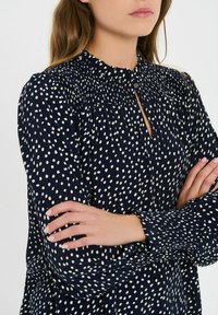 Tumed blouse valgete täppidega, kerge kangas, kogutud kaelus, nööbid ees, pikad varrukad volangidega mansettides.