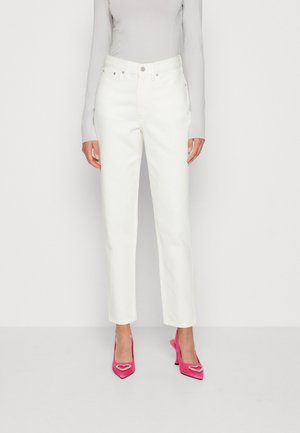 Femme portant un jean blanc droit associé à des chaussures à talons hauts rose vif ornées d'embellissements en forme de cœur.