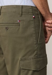 Pantalons cargo vert olive avec une finition texturée, dotés de deux poches, d'une fermeture à bouton et de petites accents brodés en forme de drapeau.