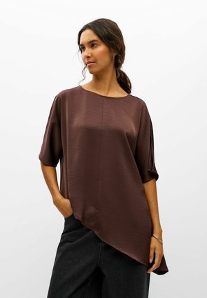 Un mannequin aux cheveux foncés attachés en arrière porte un chemisier marron ample et asymétrique ainsi qu’un pantalon noir, sur un fond blanc uni.