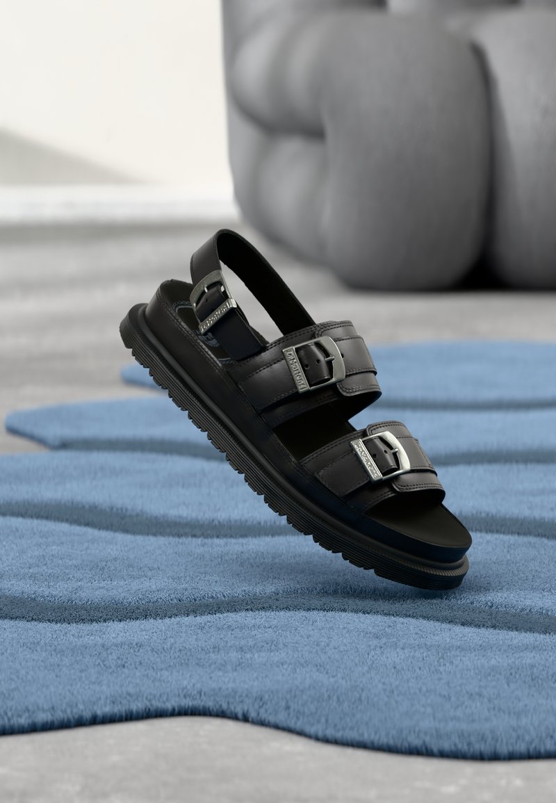 Sandalo in pelle nera con due fibbie d'argento e cinturino posteriore, posizionato sopra una superficie blu chiaro texturizzata con uno sfondo sfocato.