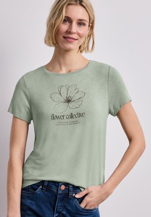 Kobieta z krótkimi blond włosami, ubrana w jasnorozświetloną zieloną koszulkę z czarnym kwiatowym szkicem i napisem „flower collective”, w połączeniu z ciemnoniebieskimi dżinsami.