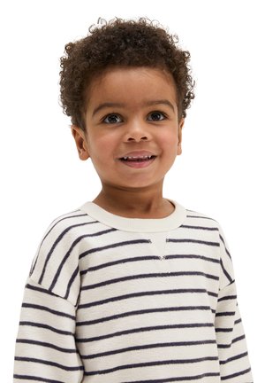 Junges Kind mit lockigem Haar, das einen weißen Pullover mit schwarzen horizontalen Streifen trägt, lächelt und schaut leicht zur Seite.