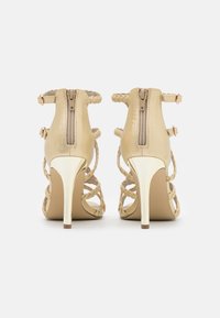 Beige korkokengät joissa on punottuja hihnoja, takana vetoketjut ja metalliset yksityiskohdat koroissa, sekä sileä pinta ja avoin muotoilu.