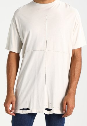 Mann trägt ein übergroßes weißes T-Shirt mit vertikalen und horizontalen Nahtdetails und kleinen Löchern nahe am Saum, kombiniert mit blauen Jeans.