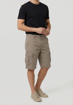 Uomo che indossa una maglietta nera, pantaloncini cargo beige con tasche laterali e sneakers beige casual, in piedi davanti a uno sfondo semplice.