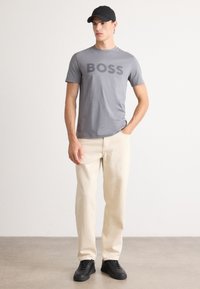 Camiseta gris de manga corta con "BOSS" impreso en el frente, combinada con pantalones rectos color crema claros y zapatillas negras. Lleva puesta una gorra negra.