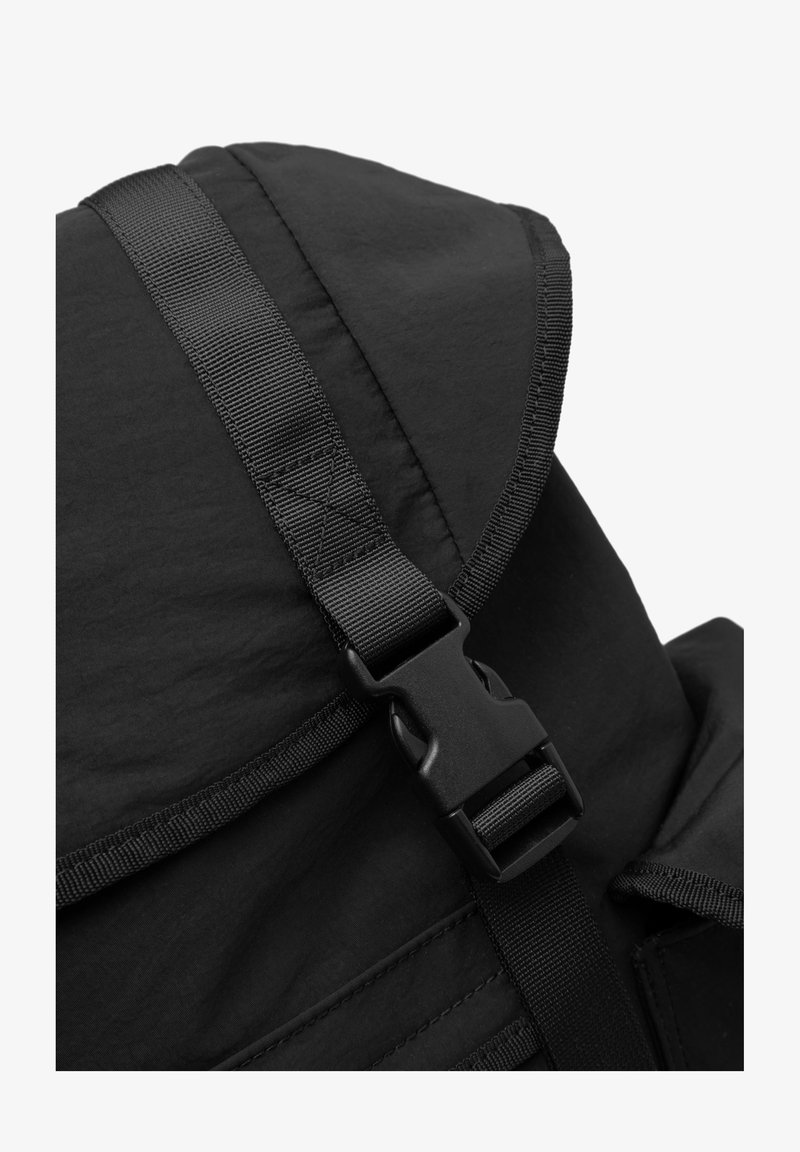 Sac à dos noir avec un dessus repliable, présentant une surface texturée, une sangle en toile réglable et une fermeture à boucle en plastique.
