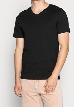 Mann trägt ein schlichtes schwarzes kurzärmeliges V-Ausschnitt-T-Shirt und hellfarbene Cordhosen, steht vor einem schlichten weißen Hintergrund.