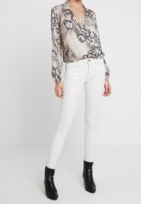 Donna che indossa una blusa con stampa serpente, jeans skinny bianchi, stivaletti neri con tacco e collane a più strati, in piedi davanti a uno sfondo bianco.